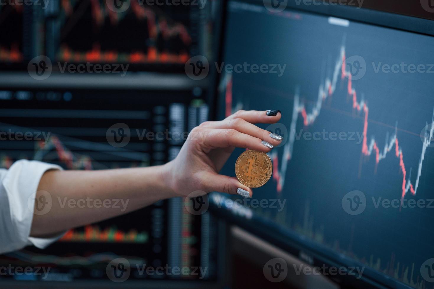 Close up view. Woman's hand holding bitcoin in modern office with many monitors with graphs photo