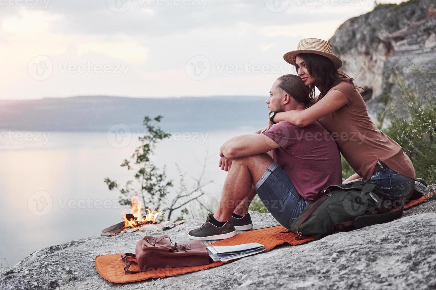 Hugging couple with backpack sitting near the fire on top of mountain enjoying view coast a river or lake. Traveling along mountains and coast, freedom and active lifestyle concept photo