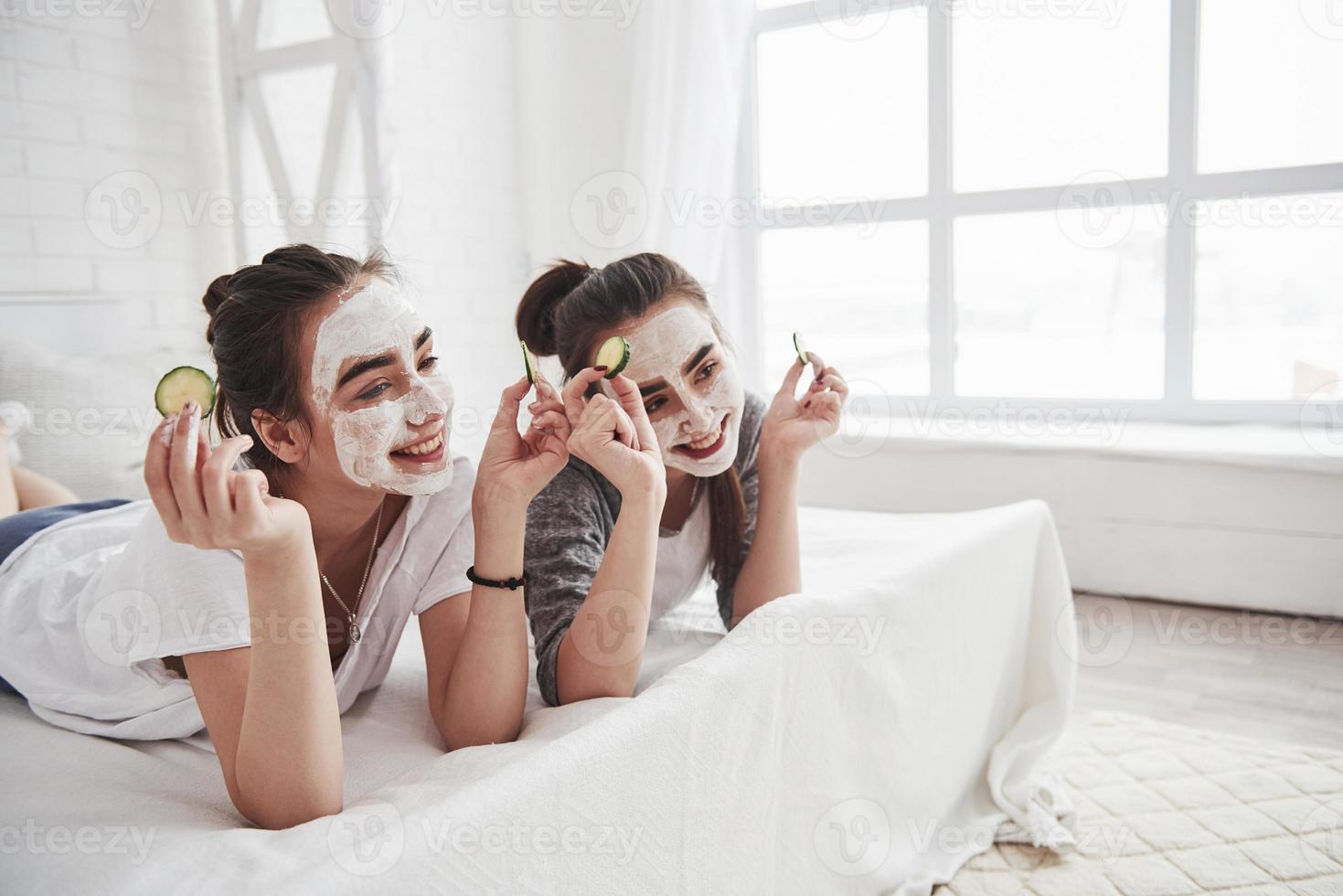 Posing for the cameramen. Conception of skin care by using white mask and cucumbers on the face. Two female sisters have weekend at bedroom photo