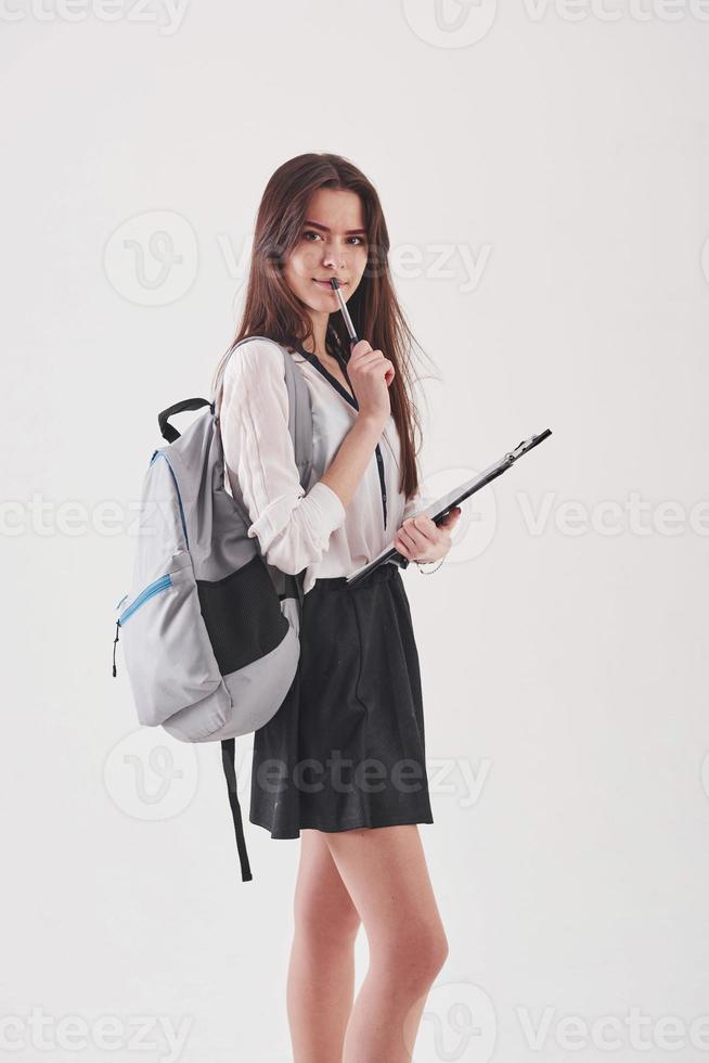 Young female student in skirt and backpack with notepad in hands stands against white background photo