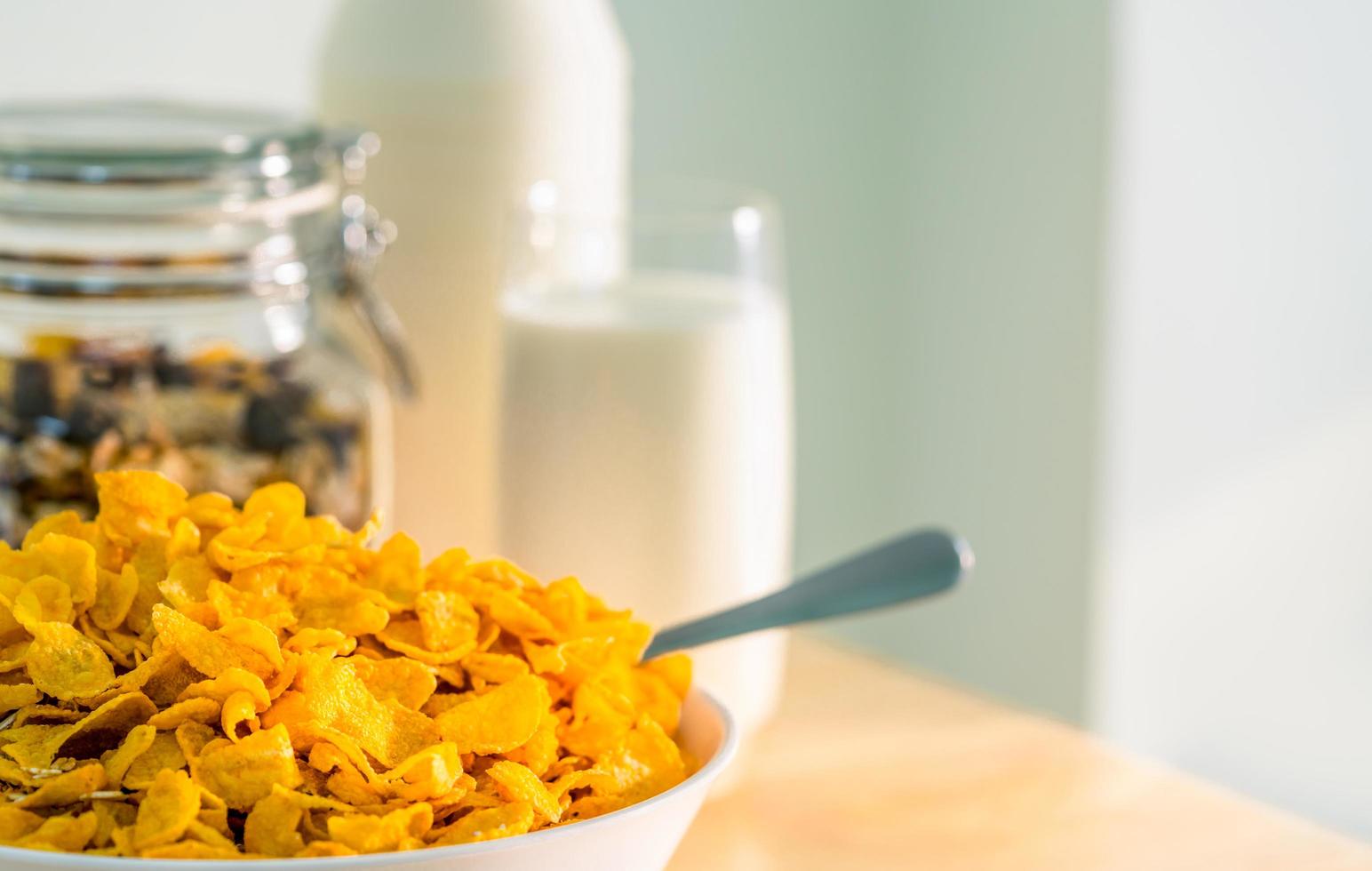 tazón de cereal con cuchara puesto sobre una mesa de madera cerca de granola en un recipiente de vidrio y un vaso de leche. desayuno de calcio para niños antes de ir a la escuela por la mañana. copos de maíz y concepto de leche foto