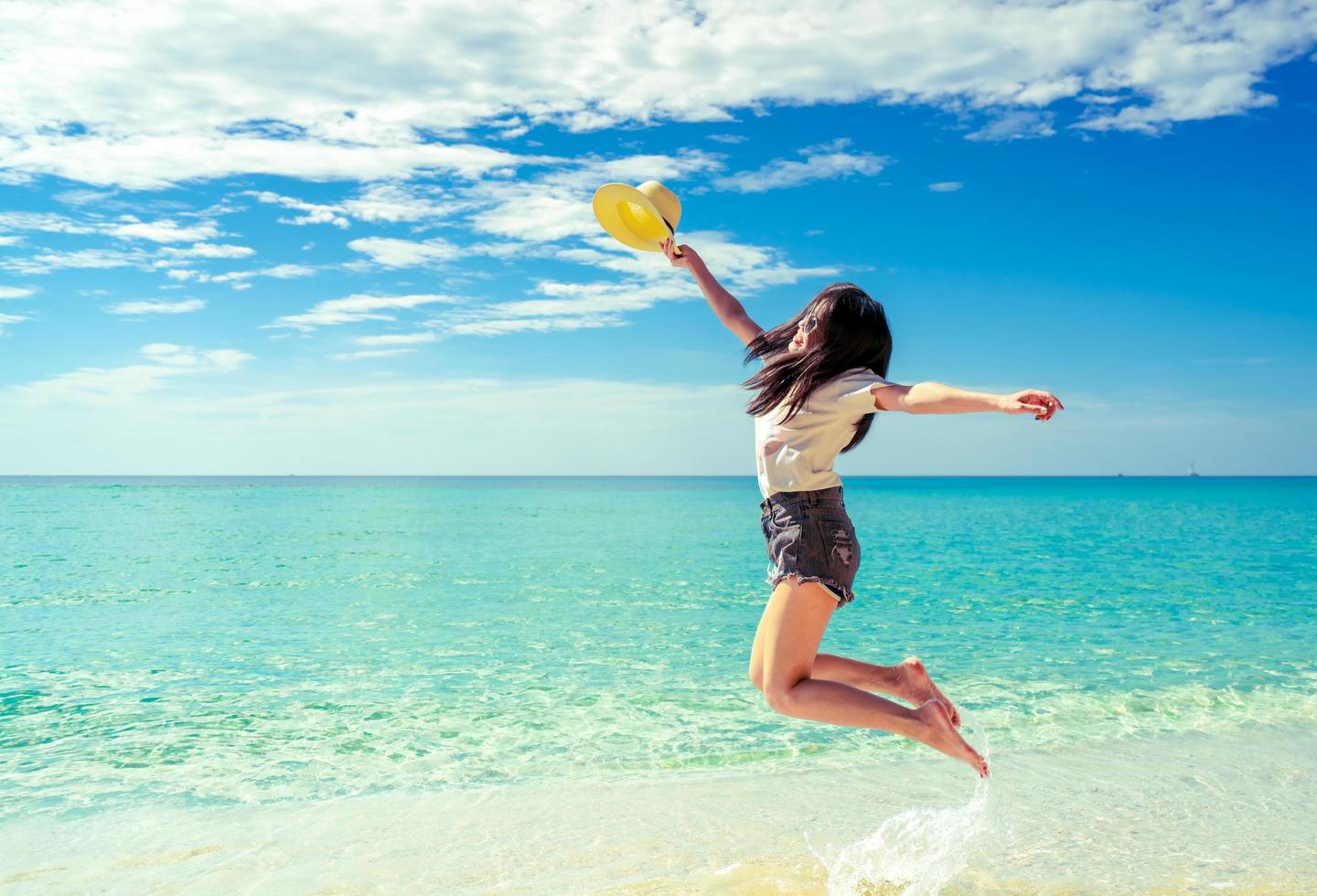 mujer joven feliz en moda de estilo casual y sombrero de paja saltando en la playa de arena. relajarse, divertirse y disfrutar de las vacaciones en la playa del paraíso tropical con cielo azul y nubes blancas. chica en vacaciones de verano. foto