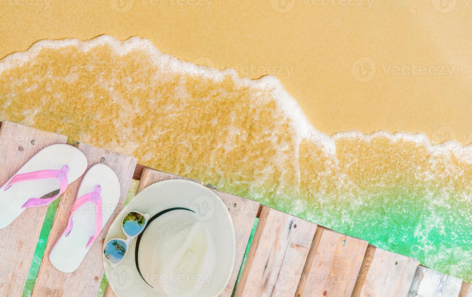 vista superior de sandalias rosadas, sombrero de paja y gafas de sol en un puente de madera sobre agua de mar verde esmeralda y playa de arena dorada. viajes de vacaciones de verano. vibras de verano. sombra de cocotero en gafas de sol. foto