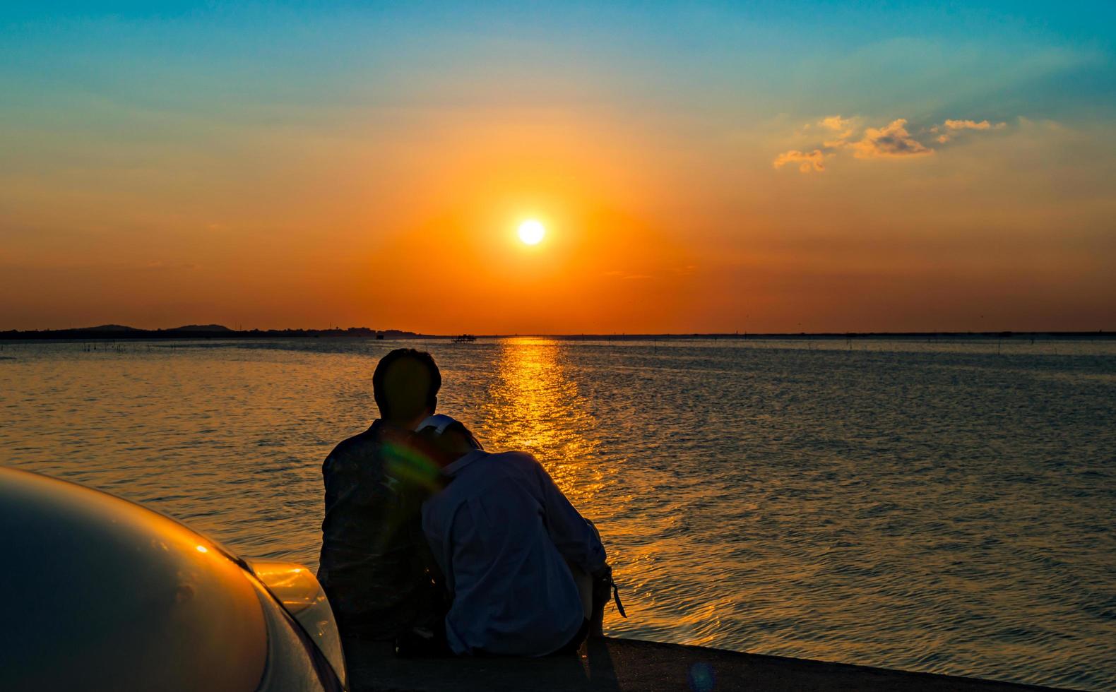 silueta de pareja feliz sentada y relajada en la playa frente al auto con cielo naranja y azul al atardecer. vacaciones de verano y concepto de viaje. pareja joven romántica saliendo en la playa. foto