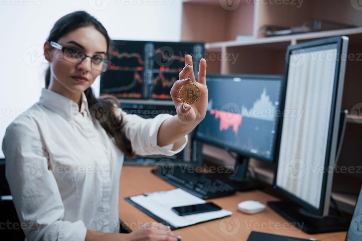 Young freelancer in eyewear. Beautiful woman holding bitcoin in hands while sitting in modern office with many monitors with graphs photo