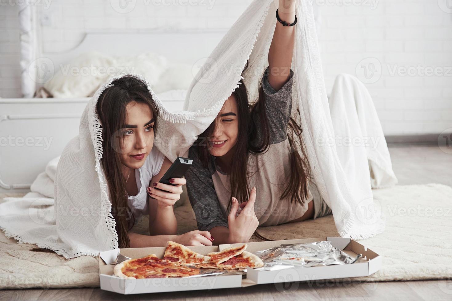 jugando con una manta caliente. hermanas comiendo pizza cuando ven la televisión mientras están acostadas en el piso de un hermoso dormitorio durante el día foto