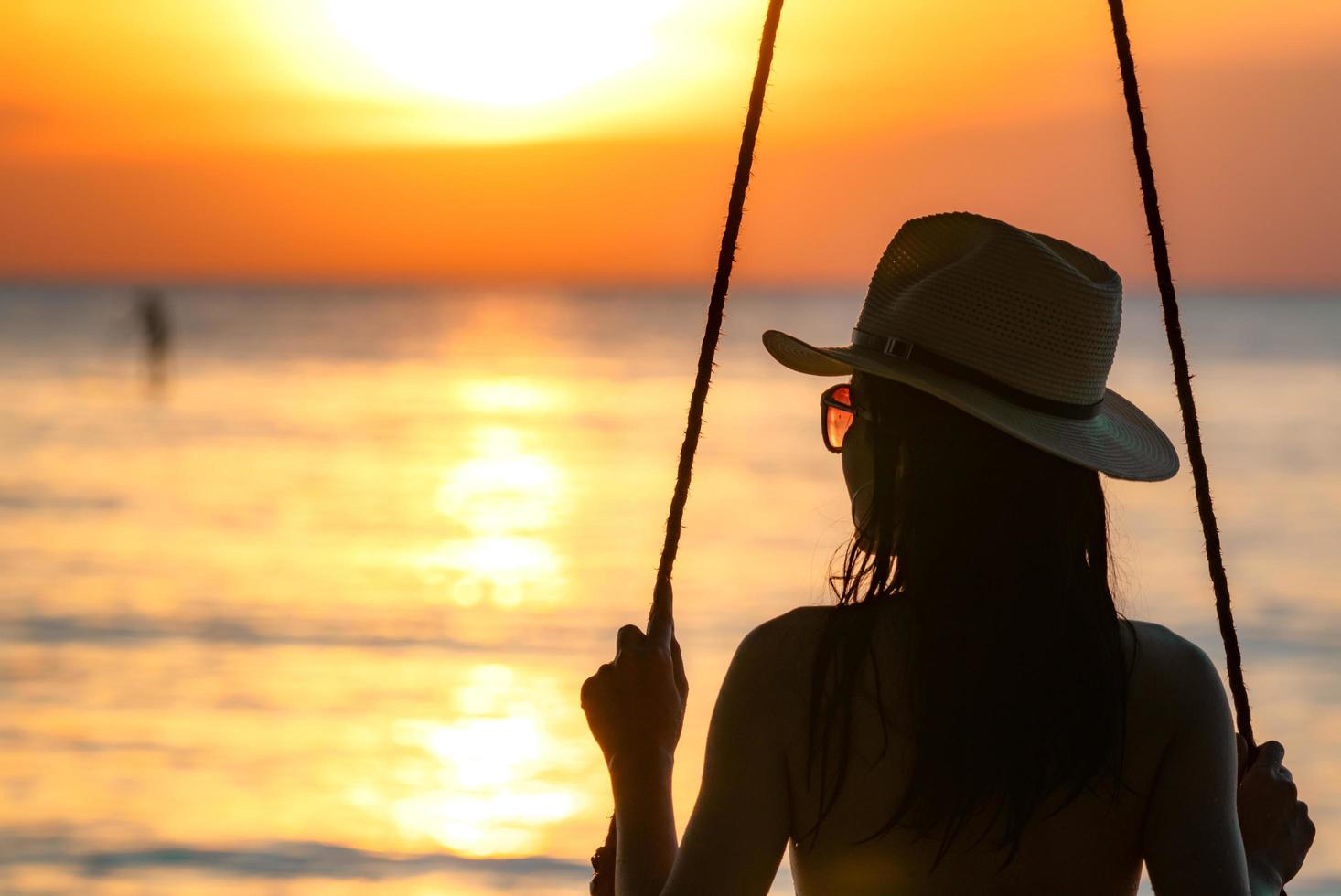 silueta mujer usa bikini y sombrero de paja columpios en la playa en vacaciones de verano al atardecer. chica en traje de baño se sienta en los columpios y mira la hermosa puesta de sol. vibras de verano. mujer viaja sola. foto