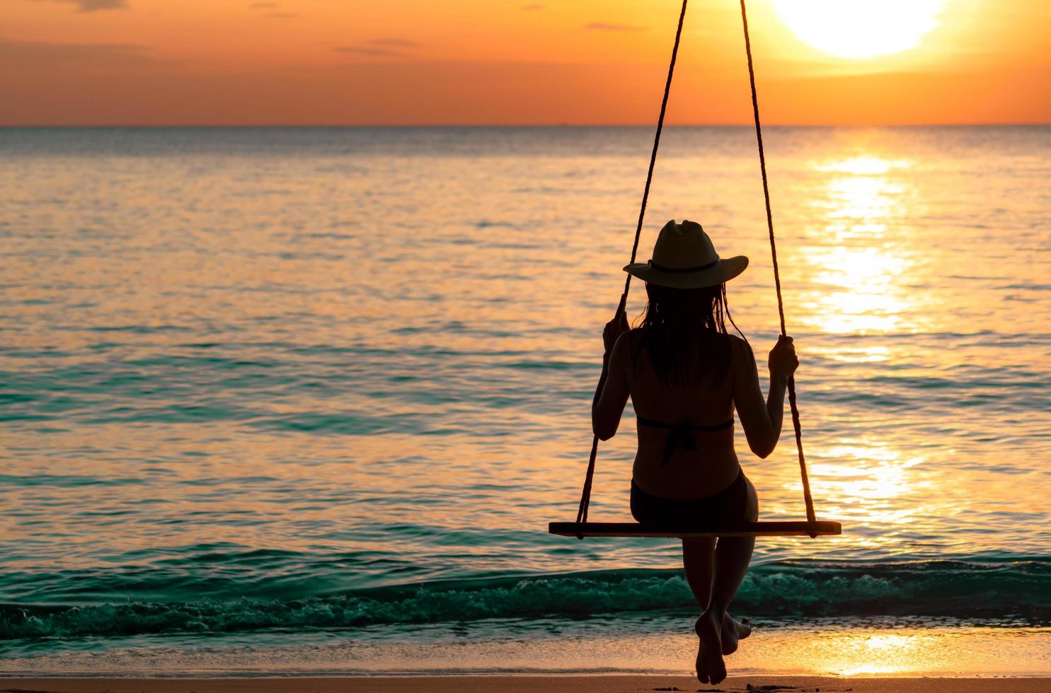 silueta mujer usa bikini y sombrero de paja columpios en la playa en vacaciones de verano al atardecer. chica en traje de baño se sienta en los columpios y mira la hermosa puesta de sol. vibras de verano. mujer viaja sola. foto