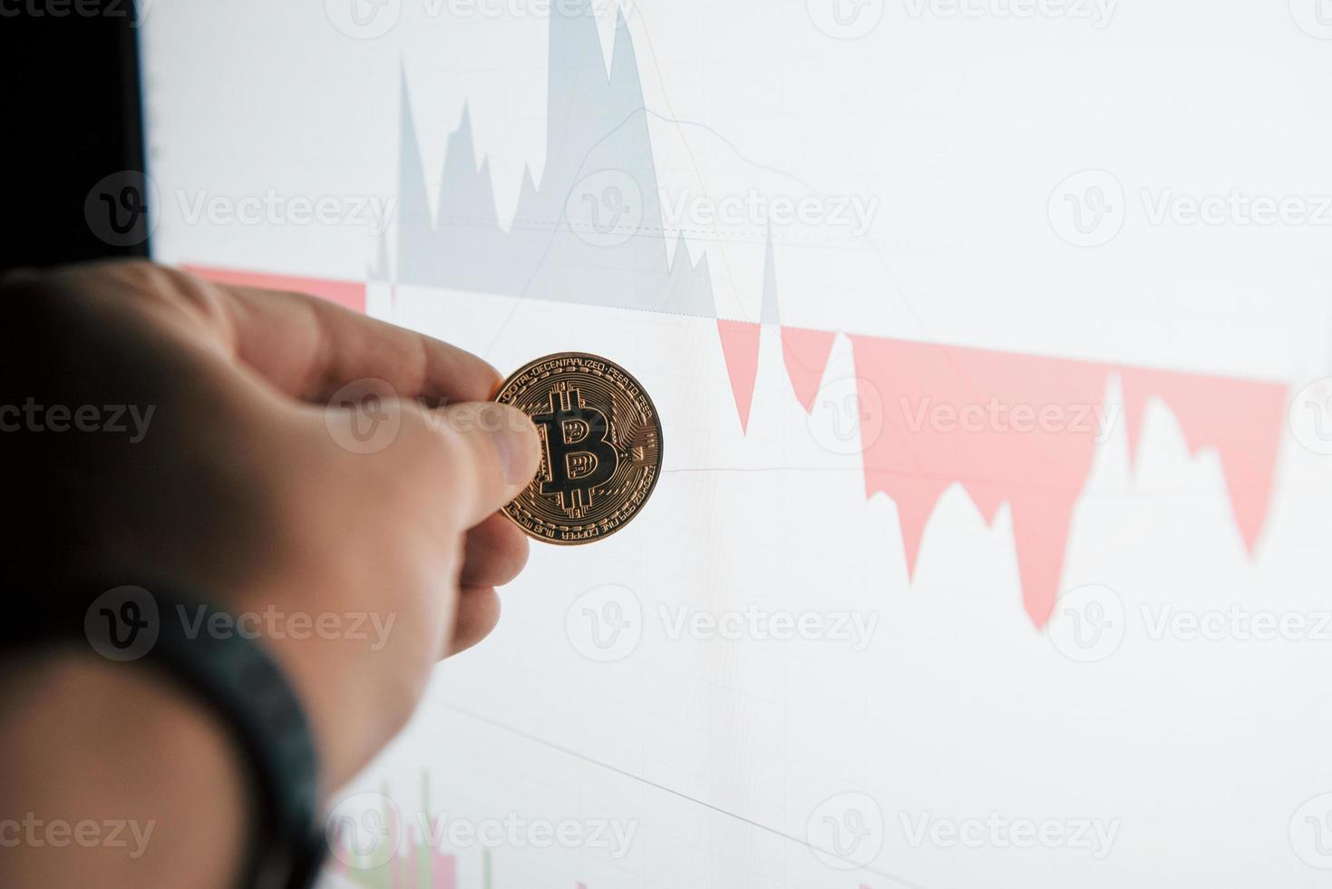 Focused photo. Man's hand holding bitcoin in modern office against monitor with graph photo