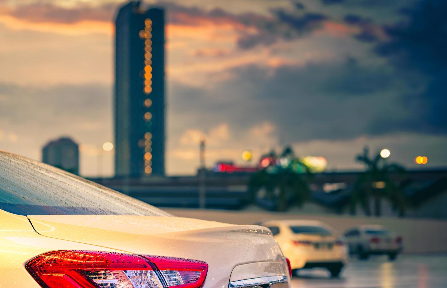 Rear view of car parked in the city near high building after rain with grey and orange sky and clouds and have water droplet on surface of car. Hybrid auto concept. Travel in the city on rainy season. photo