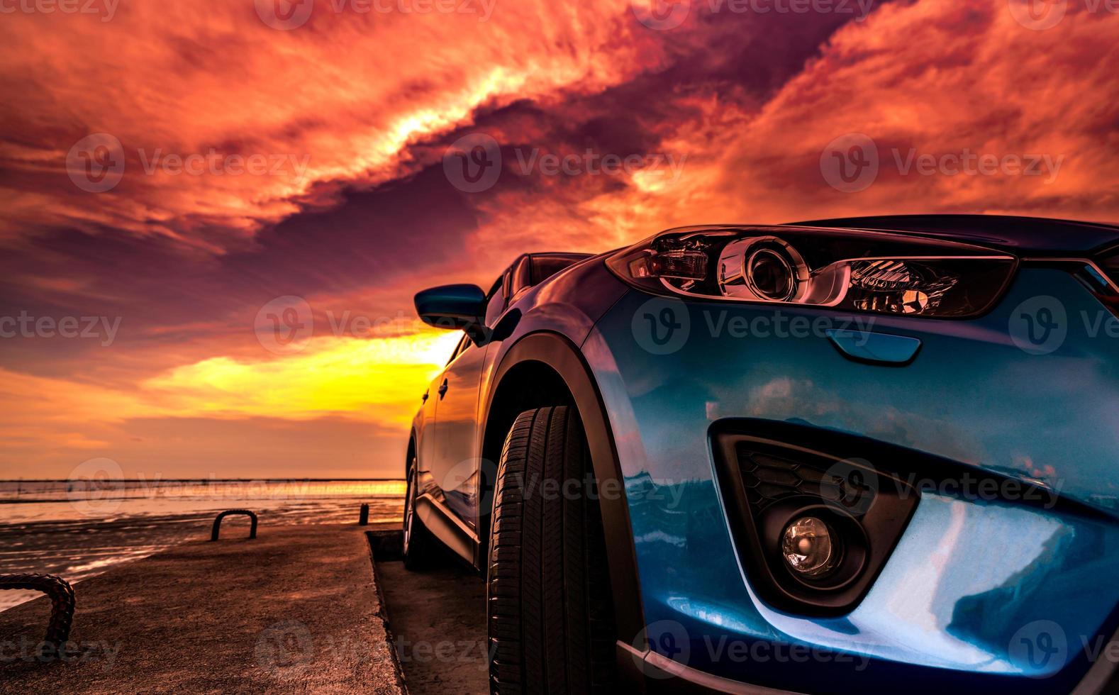 Blue compact SUV car with sport, modern, and luxury design parked on concrete road by the sea at sunset. Front view of beautiful hybrid car. Driving with confidence. Travel on vacation at the beach. photo