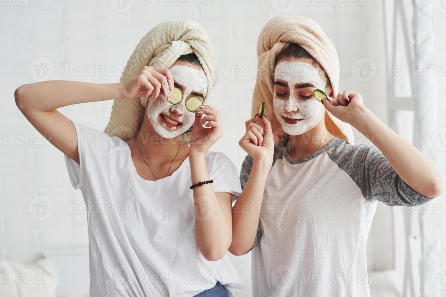 Dancing a little. Conception of skin care by using fresh cucumber rings and white mask on the face. Two female sisters have weekend at bedroom photo