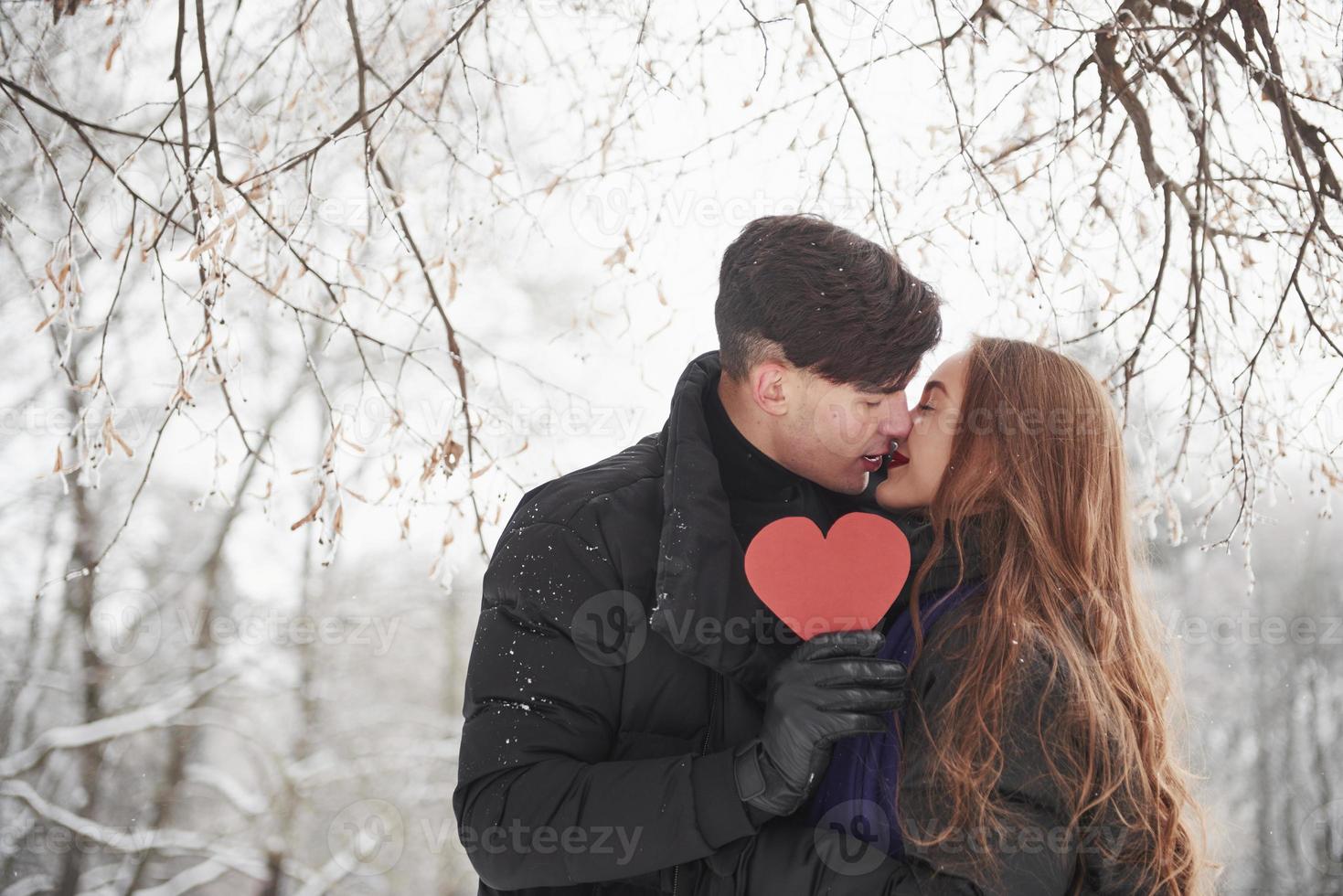 Holding red heart shaped cardboard. Gorgeous young couple have good time together in snowy forest photo