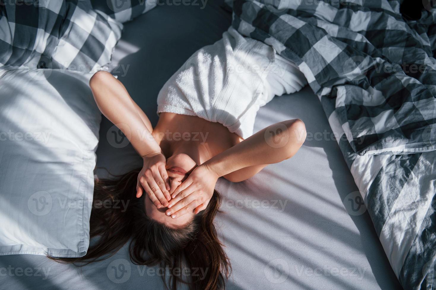 Gorgeous girl covered her face with hands. Pretty young woman lying on the bed at morning time in her room photo