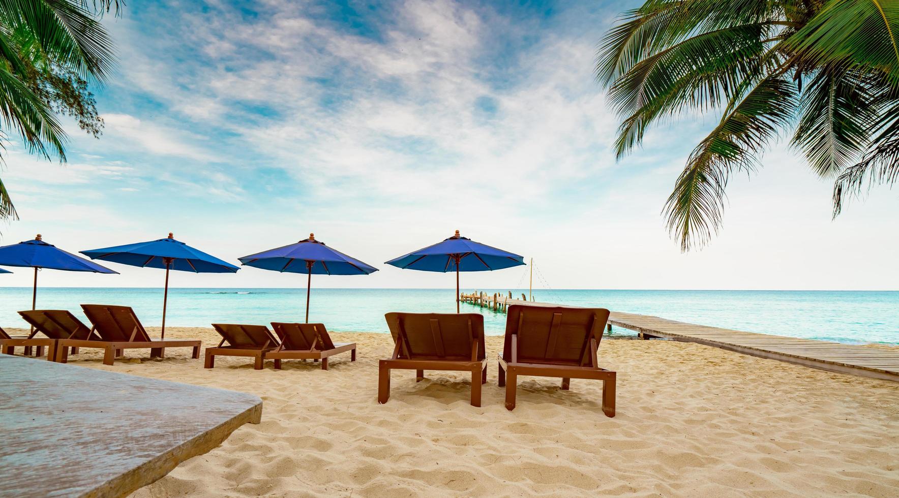 silla de playa de madera y sombrilla en la playa de arena del resort al lado del puente. viajes de vacaciones de verano. viajes de vacaciones tropicales. tres parejas de tumbonas y cocoteros con cielo azul y nubes blancas. foto