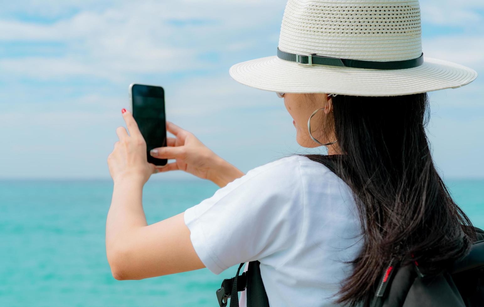 Young Asian backpacker woman wear hat use smartphone taking selfie at pier. Summer vacation at tropical paradise beach. Happy hipster girl travel on holiday. Woman enjoy and relax life. Summer vibes. photo