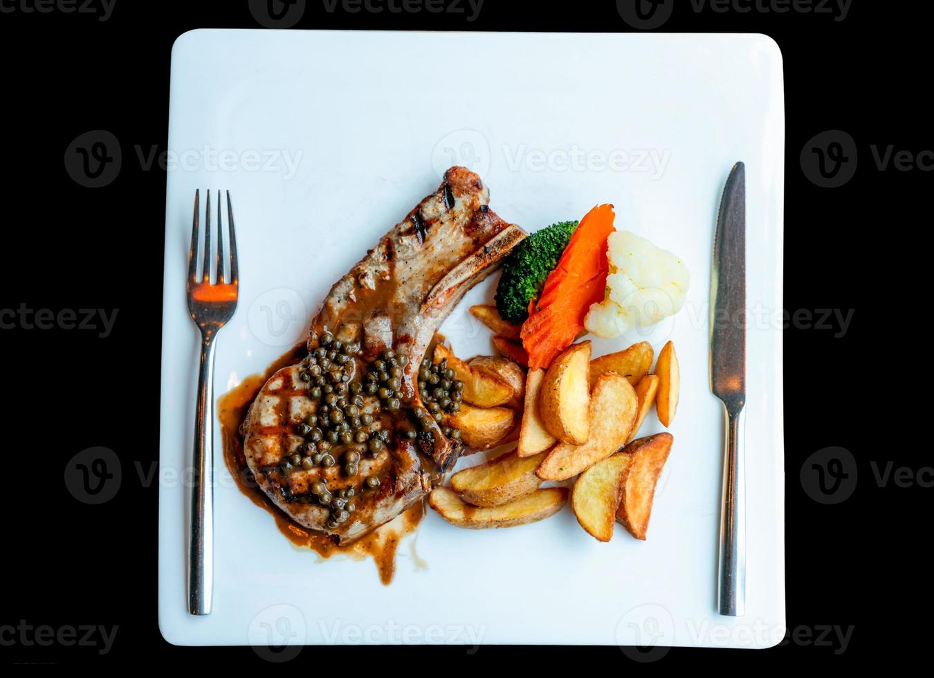 Top view of steak topped with sauce and pepper serve with potato fried and carrots, cauliflower and broccoli steamed. Steak on white plate with stainless steel fork and knife. Delicious protein food. photo