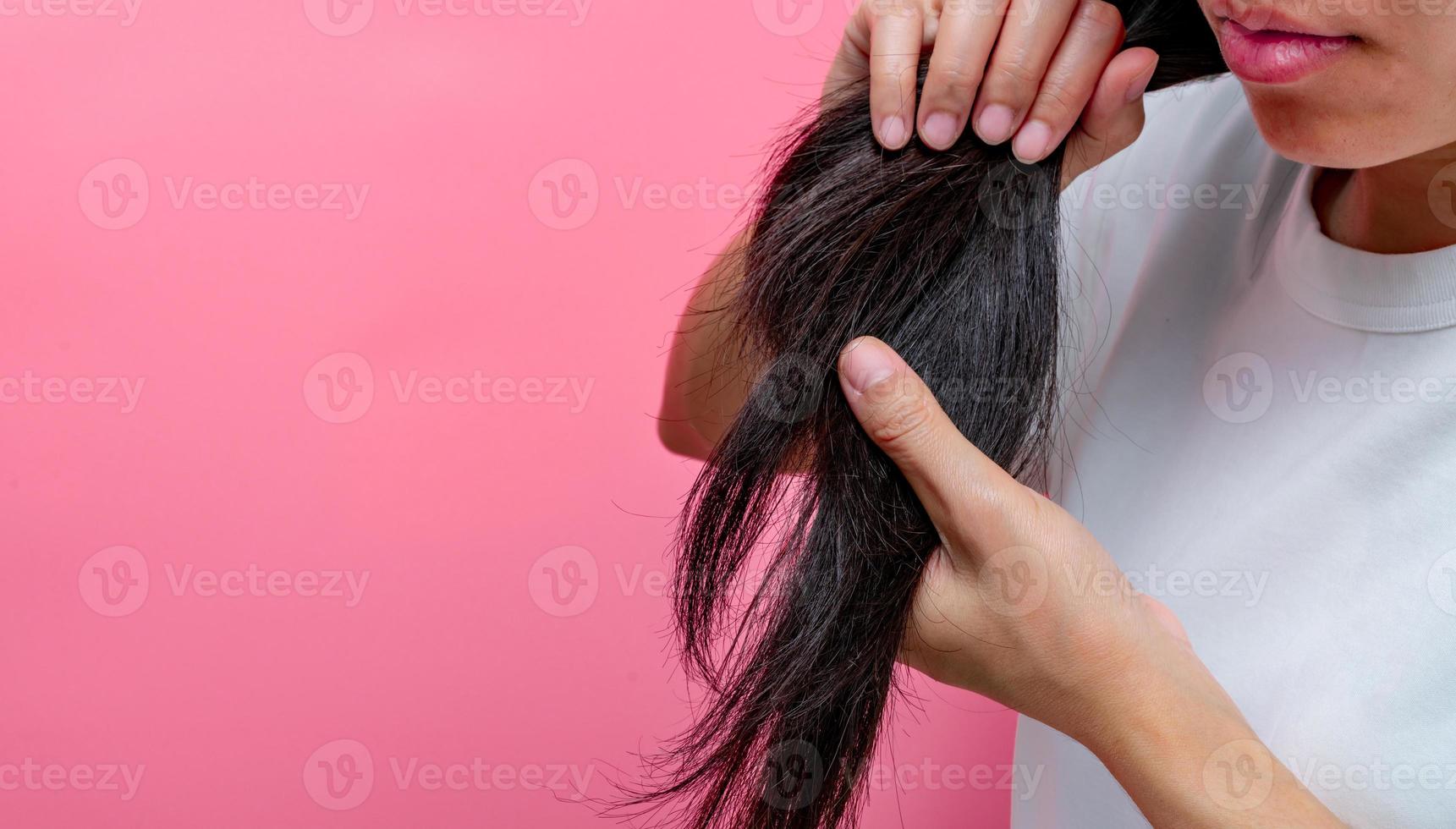 mujer asiática con labios secos sosteniendo el cabello dañado sobre fondo rosa. El cabello largo, negro, seco y quebradizo necesita champú y acondicionador para un tratamiento de spa. pérdida de cabello y problemas de cabello fino en las mujeres. foto