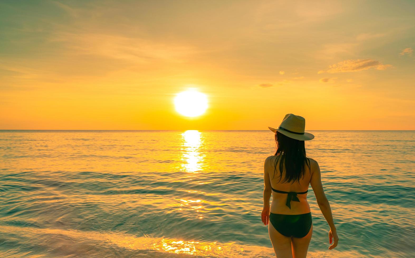 Silhouette adult woman walking at tropical sea with beautiful sunset sky at paradise beach. Happy girl wear bikini and straw hat relaxing summer vacation. Holiday travel. Summer vibes. Life goes on. photo