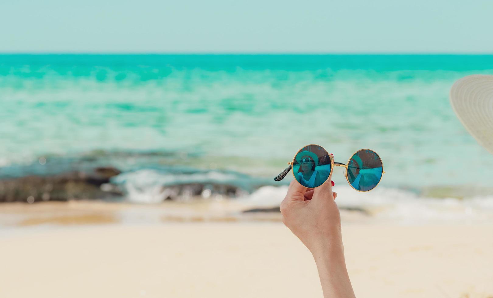 Woman hand holding sunglasses and sit on sand beach. Reflection of woman in sunglasses. Women wear straw hat relax at tropical beach on summer vacation. Sunny day on holiday. Travel alone on summer. photo