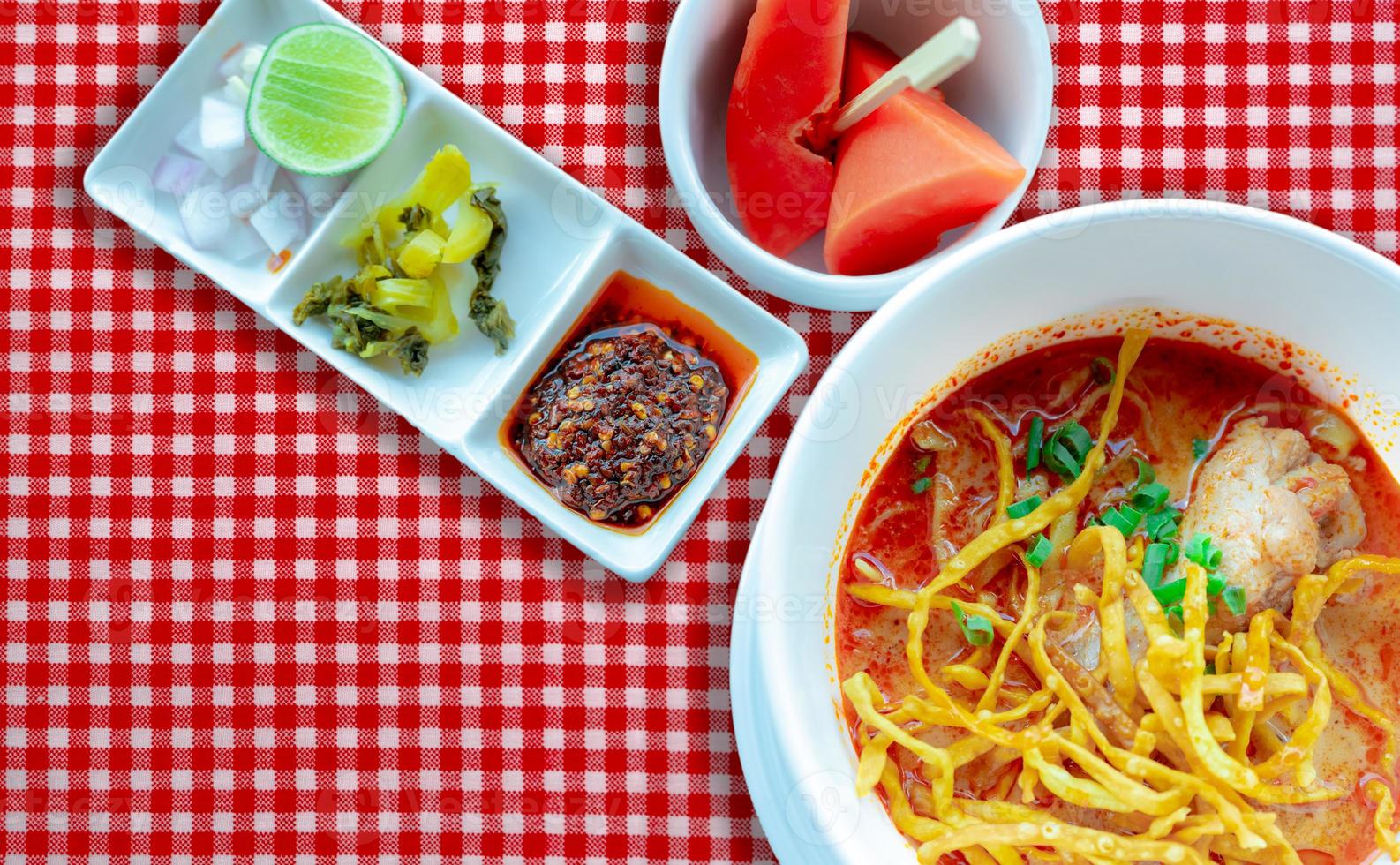 Northern Thai curry noodle soup with chicken and side dish in white bowl on red checkered tablecloth. Top view of Khao Soi and ripe papaya with wooden fork in bowl. Thai traditional food menu. photo