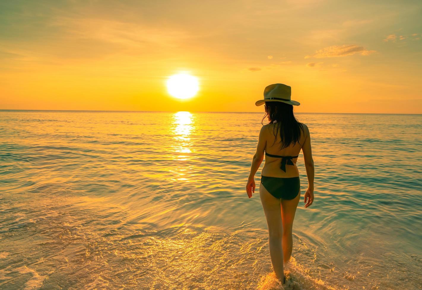 Silhouette adult woman walking at tropical sea with beautiful sunset sky at paradise beach. Happy girl wear bikini and straw hat relaxing summer vacation. Holiday travel. Summer vibes. Life goes on. photo