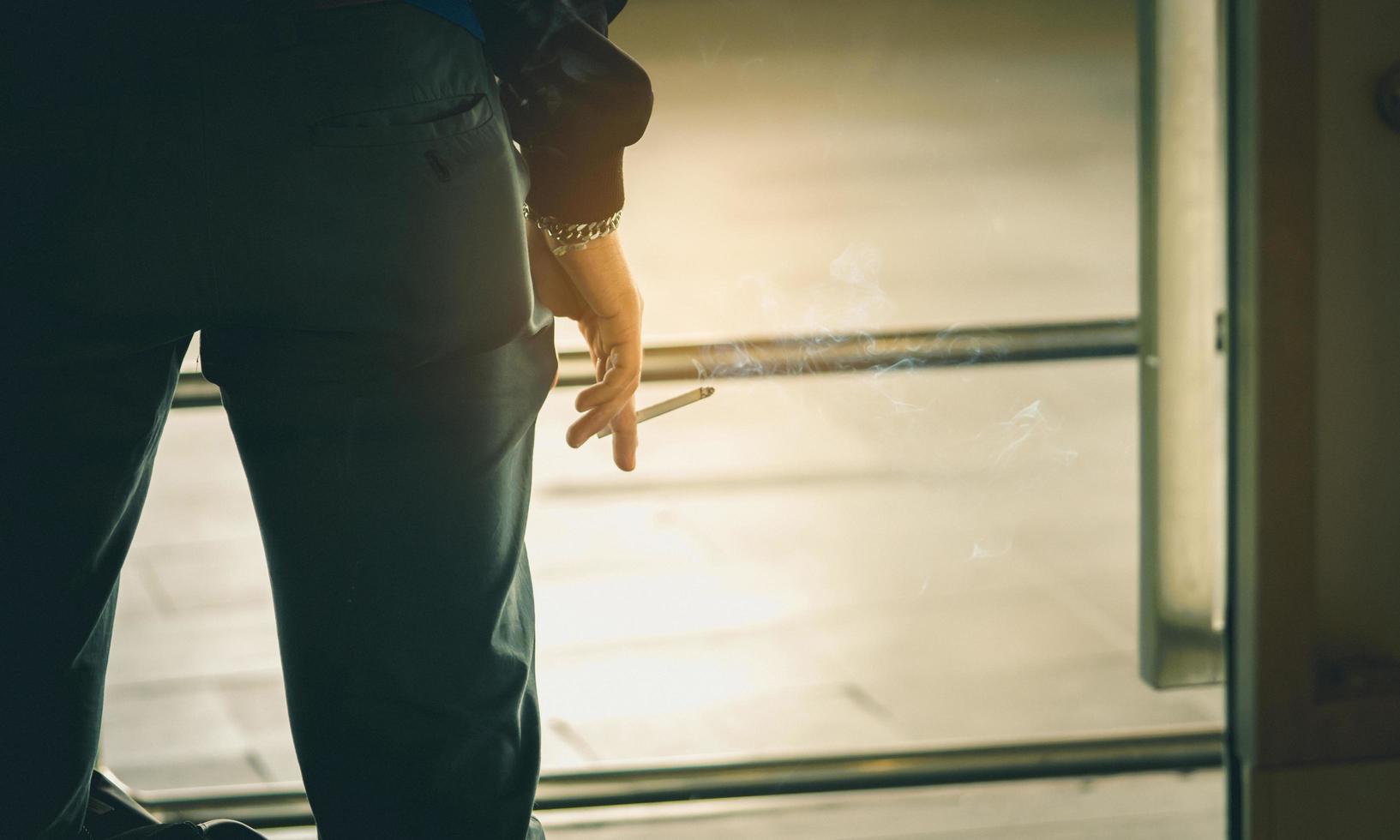 vista trasera de primer plano de un hombre asiático con pantalones y camisa de manga larga. hombre mano sosteniendo cigarrillo y humo. hombres de negocios fumando cigarrillos en la oficina cuando estaba estresado por el trabajo. adicto a la nicotina foto