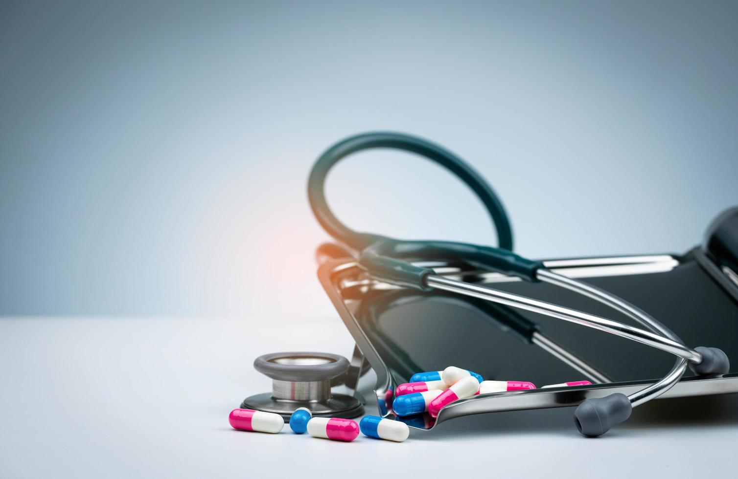 Green stethoscope with pile of antibiotic capsule pills on drug tray and spread on white table. Antimicrobial drug resistance and overuse. Medical equipment for doctor. Clinical Cardiology concept. photo