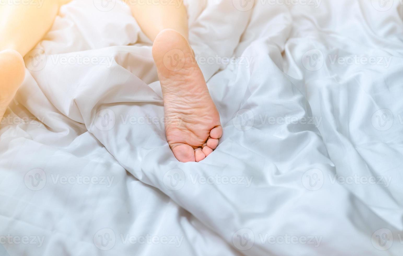 Cierra los pies descalzos de la mujer en la cama sobre una manta blanca y una sábana en el dormitorio de la casa o del hotel. concepto de dormir y relajarse. mañana perezosa. descalzo de mujer acostada en cama blanca y edredón. foto