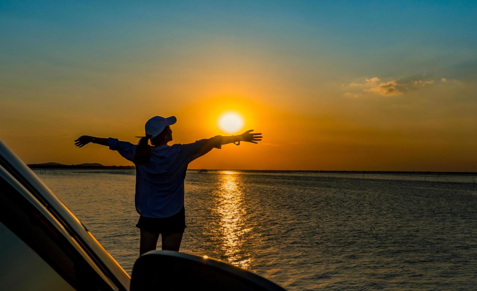 Silhouette of beautiful happy young woman wear a cap relaxing on the beach in front of the car with orange and blue sky at sunset. Summer vacation and travel concept. Environmentally friendly. photo