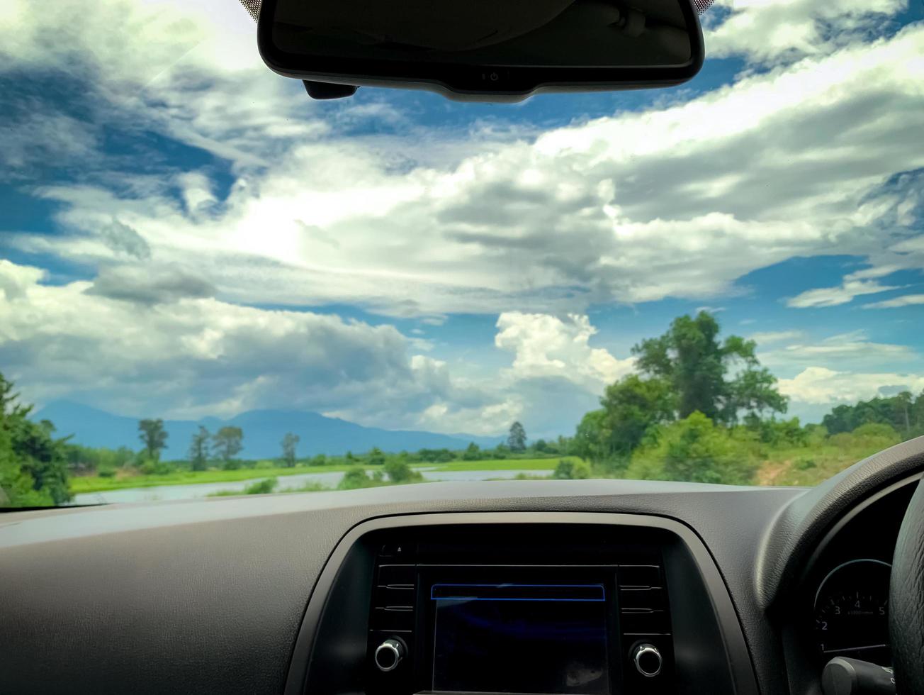 Beautiful landscape view from inside car. Steering wheel and dashboard of car interior. Road trip travel with scenic view of mountain, lake, and forest. Blue sky and white fluffy clouds. Vacation time photo