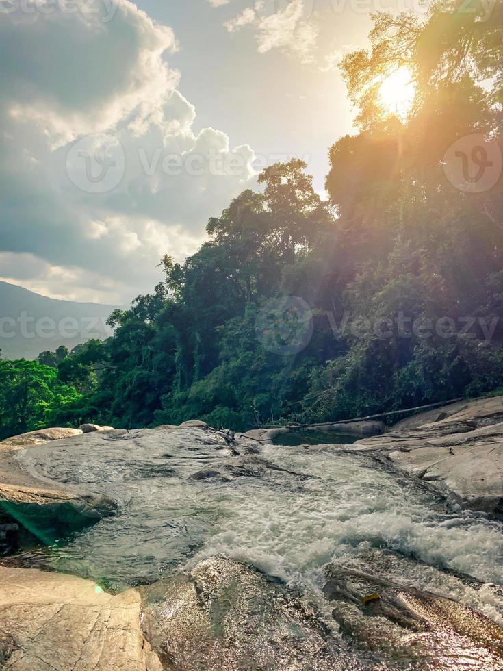Beautiful waterfall at the mountain with blue sky and white clouds and orange sun light. Waterfall in green tree forest. Waterfall is flowing. Nature background. photo
