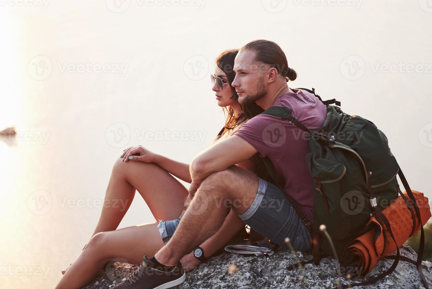 Hugging couple with backpack sitting on top of rock mountain enjoying view coast a river or lake. Traveling along mountains and coast, freedom and active lifestyle concept photo
