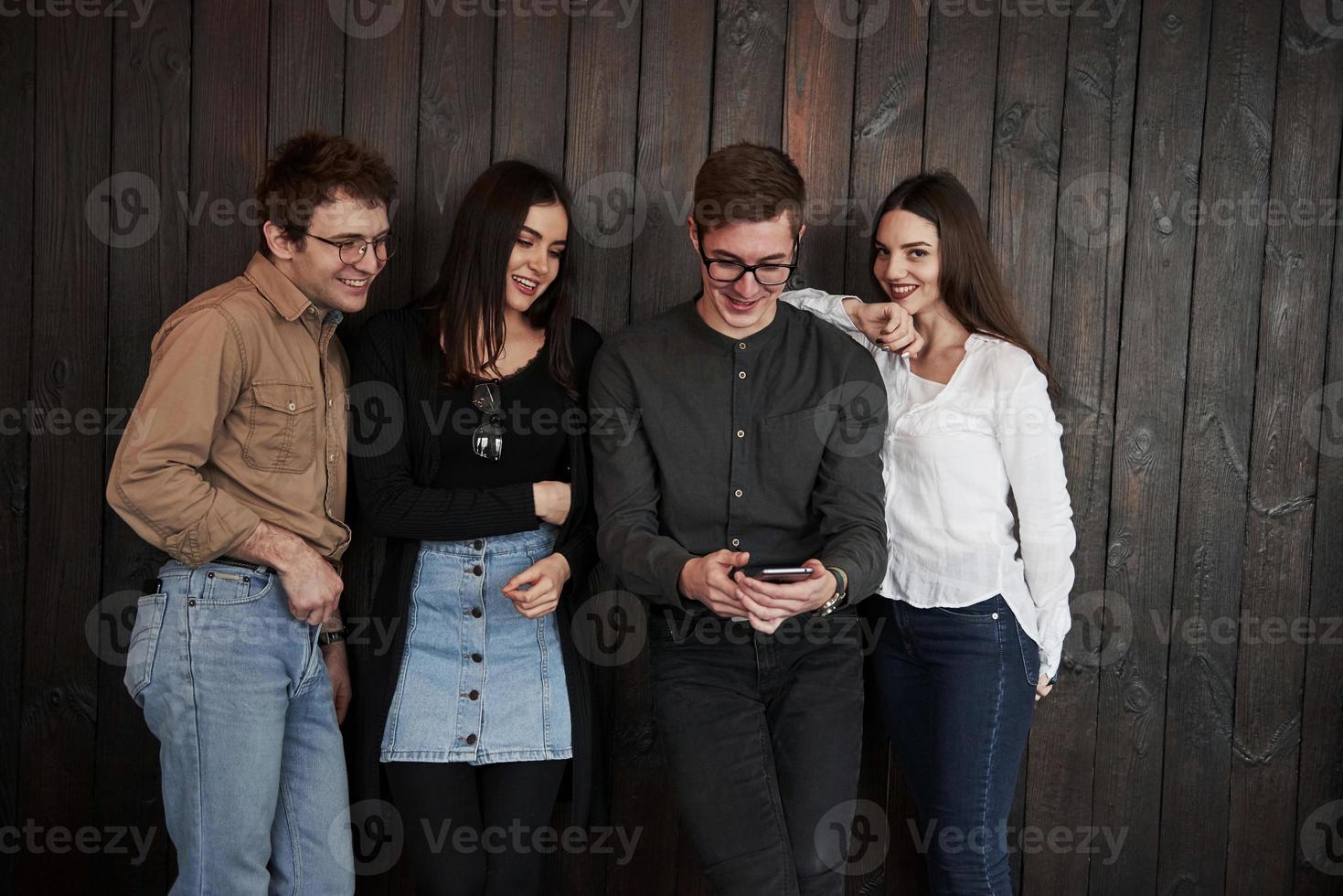 Guy sarching for some fun content. Youth stands against black wooden wall. Group of friends spending time together photo