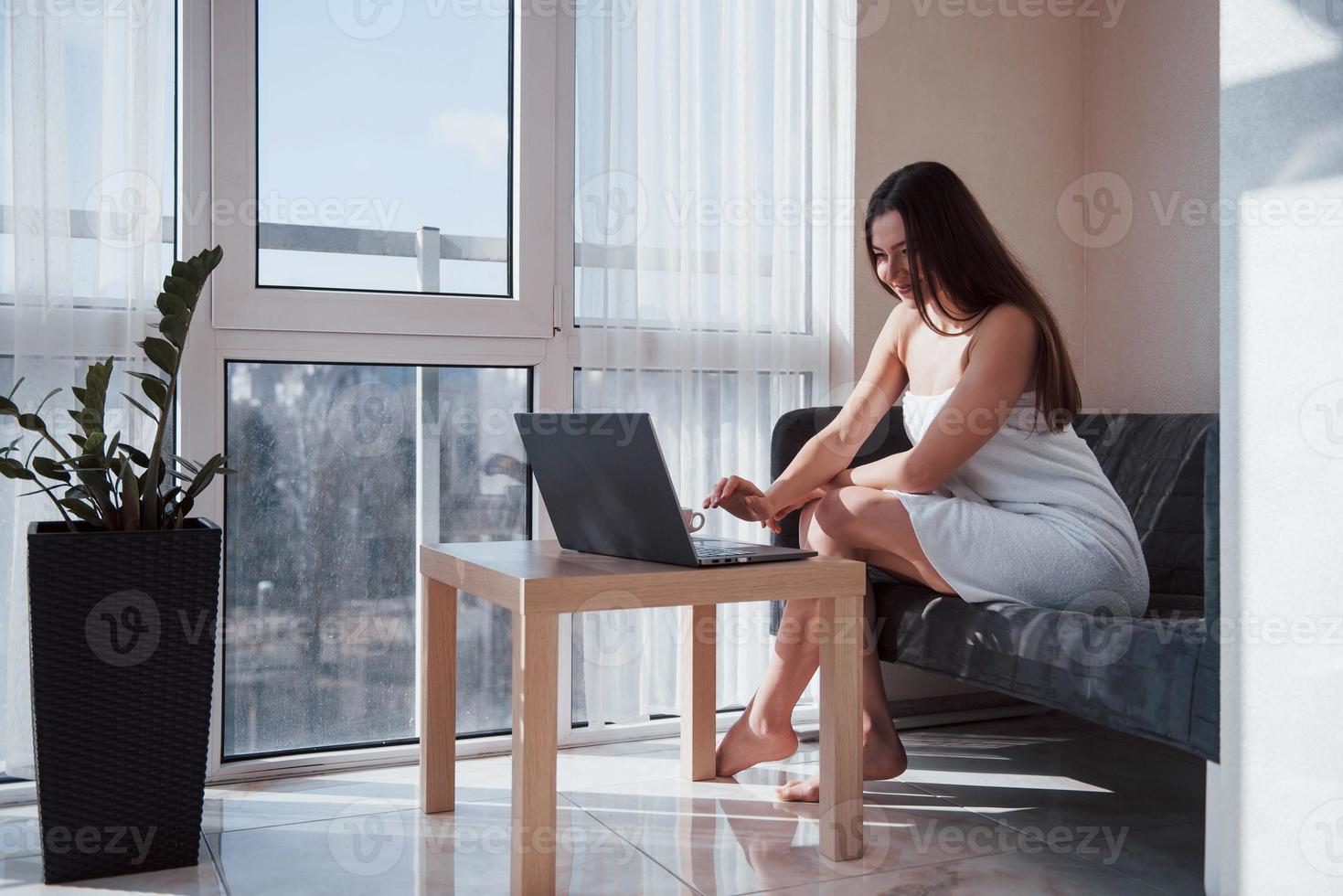 City through the window. Pretty young woman sitting on the sofa at morning time in her room with laptop photo