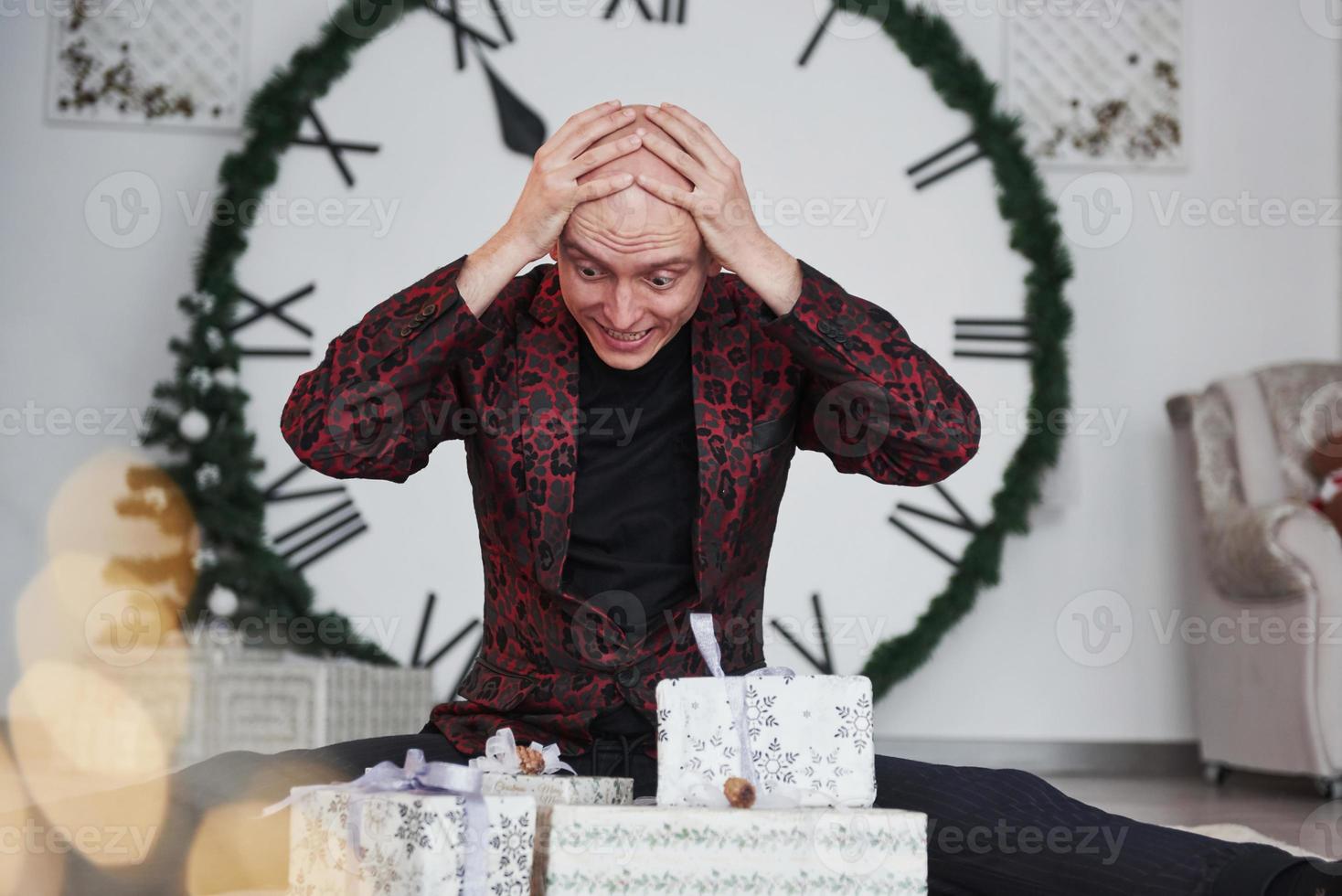 Too many presents. Man with big clock behind him sitting with New year gift boxes in holiday clothings photo