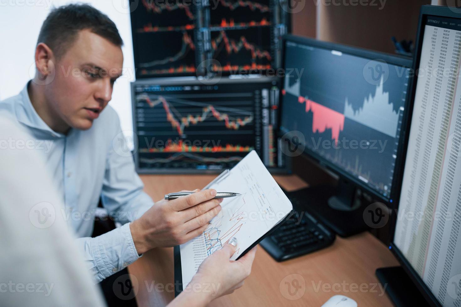 Notepad in hands with some graphs. Team of stockbrokers are having a conversation in a office with multiple display screens photo