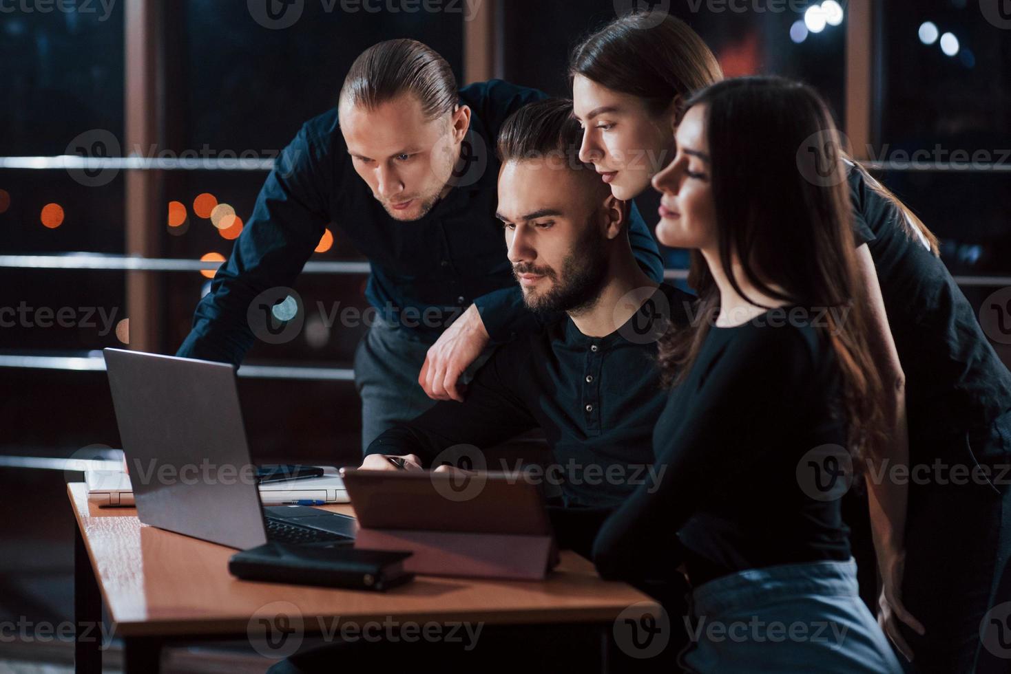 All look to the screen of the laptop. Team of young business people works on their project at night time in the office photo