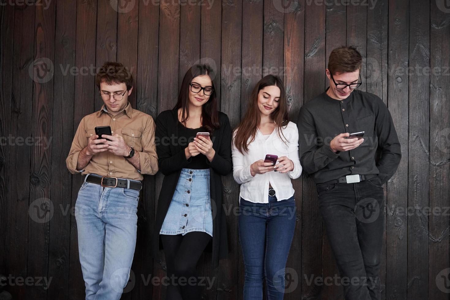 todos los adictos a su teléfono inteligente. el joven se para contra la pared de madera negra. grupo de amigos pasando tiempo juntos foto