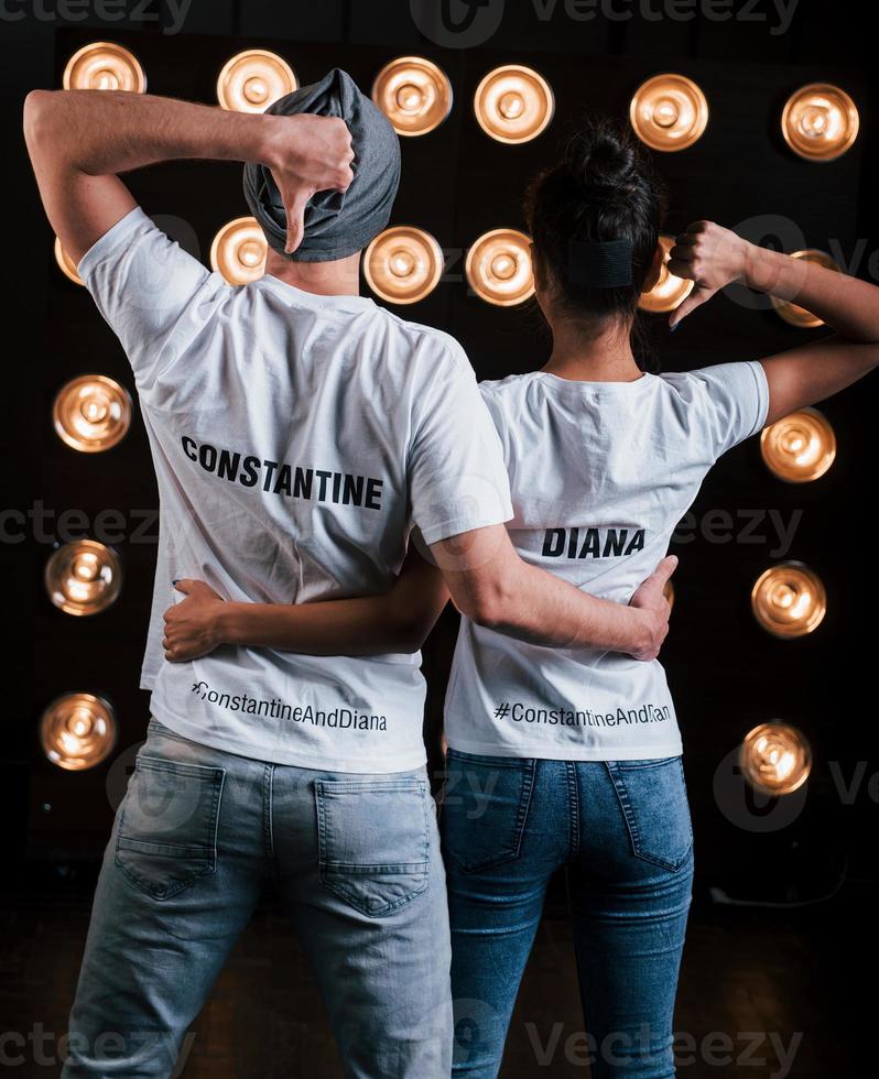 Gestures with thumbs up and hugs. Back view of couple in shirts with their names on it standing next to studio lights photo