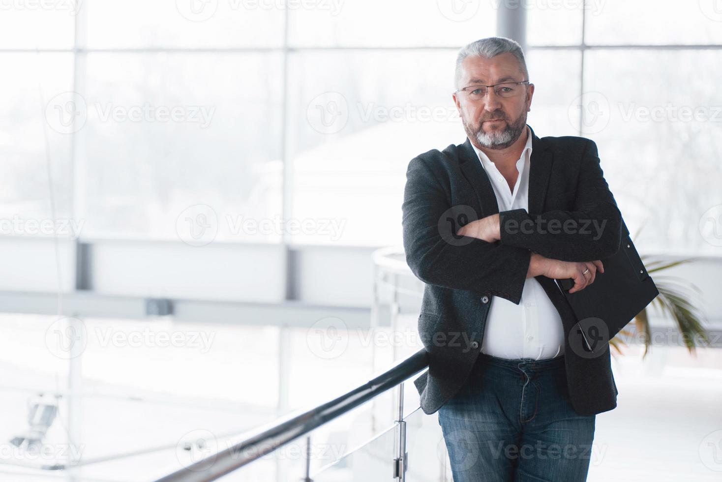 With arms crossed. Photo of senior businessman in the spacious room with plants behind. Holding and reading documents