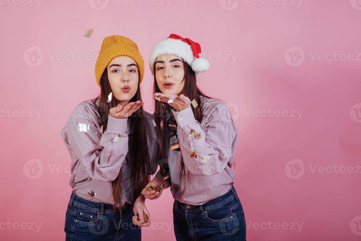 It's Christmas time. Confetti and good mood for all. Air kisses. Two sisters twins standing and posing in the studio with pink background photo