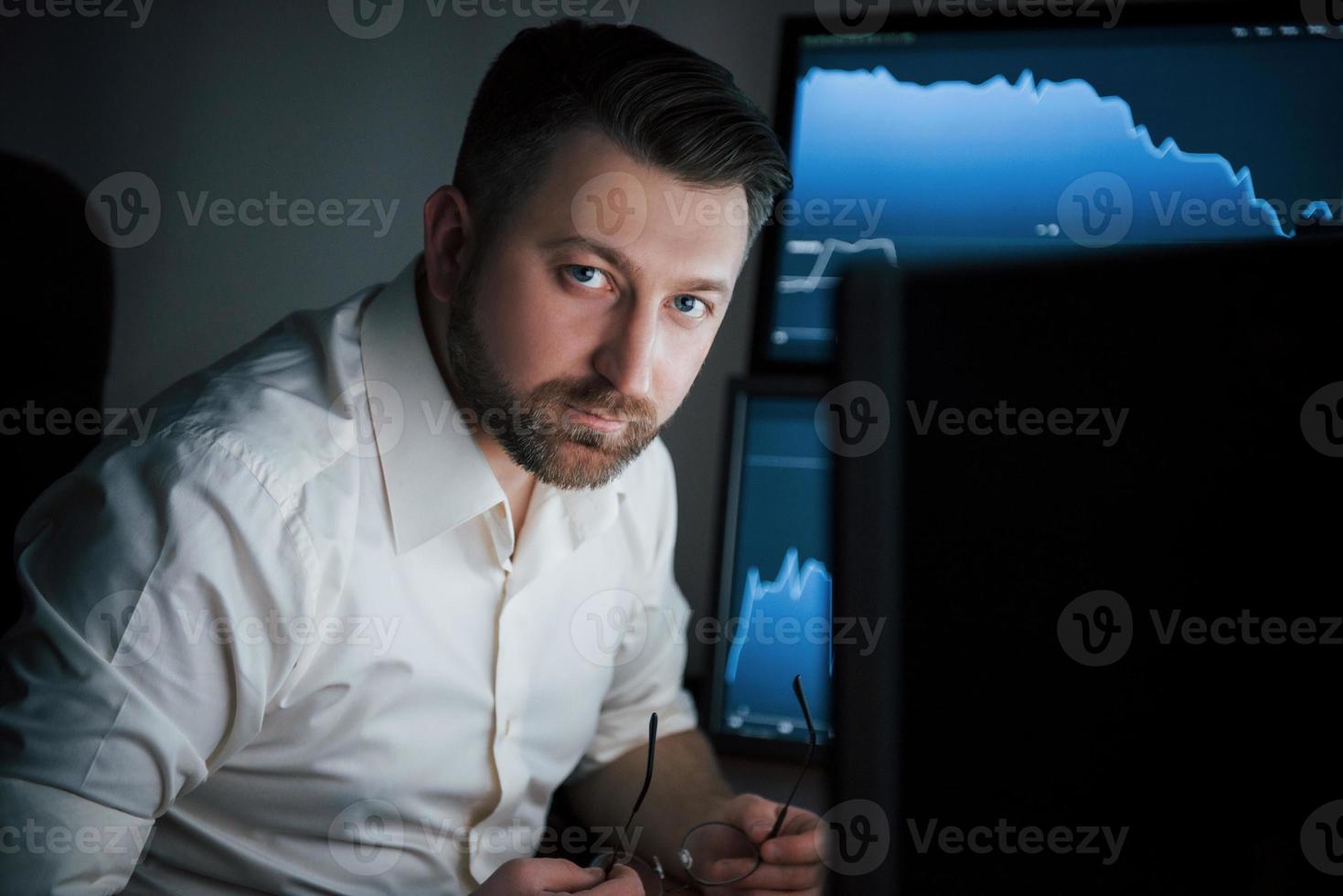 Looking into the camera. Bearded man in white shirt works in the office with multiple computer screens in index charts photo