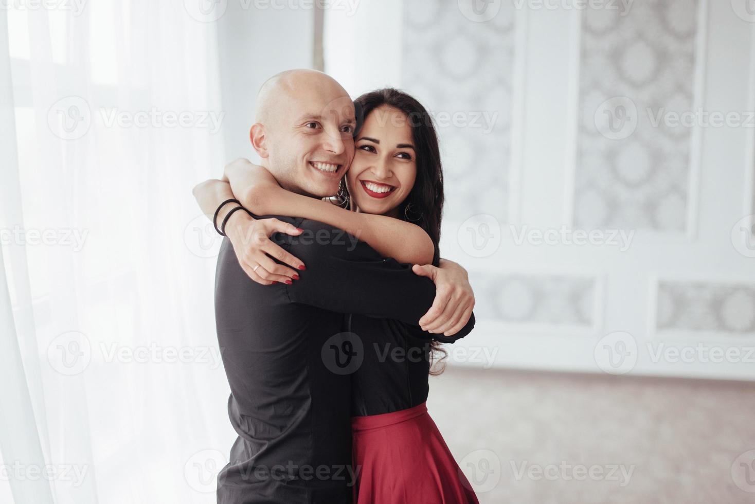 Cute and cheerful people. Hug each other and smiling. Portrait of happy couple indoors. Bald guy and brunette woman stands in the white room photo