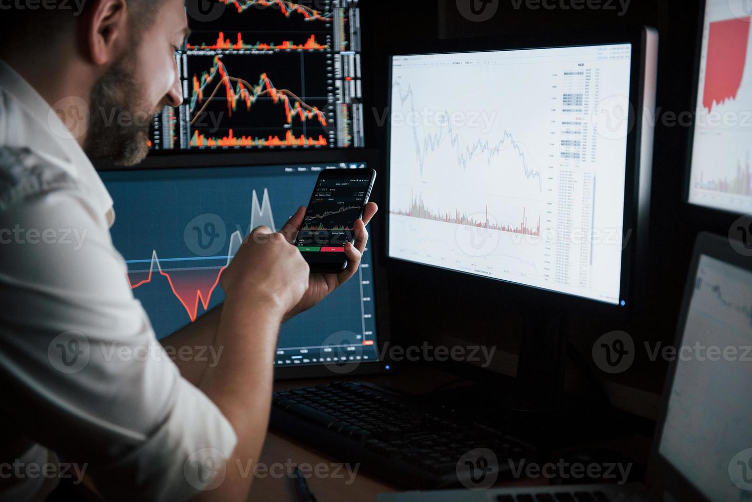 Satisfied by results. Bearded man in white shirt works by using smartphone in the office with multiple computer screens in index charts photo