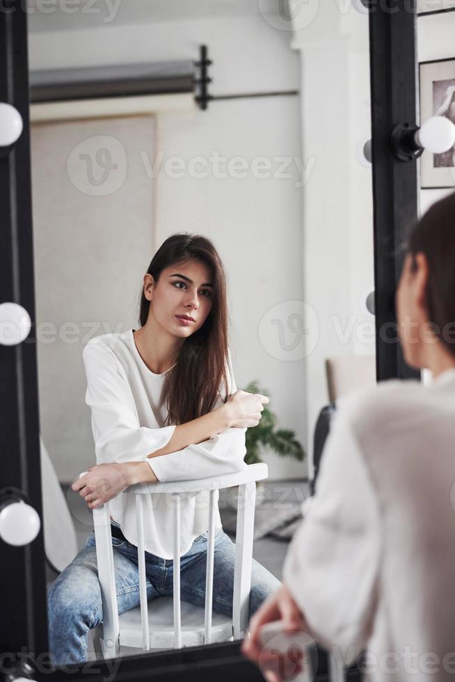 Sitting on the chair and looking in the mirror. Young brunette in the room with white walls and daylight that comes from the window photo