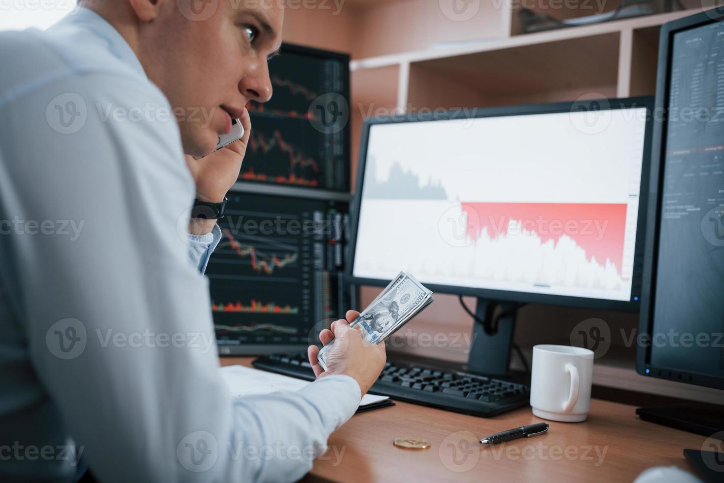 Haves conversation while holding money in the hand. Man working online in the office with multiple computer screens in index charts photo