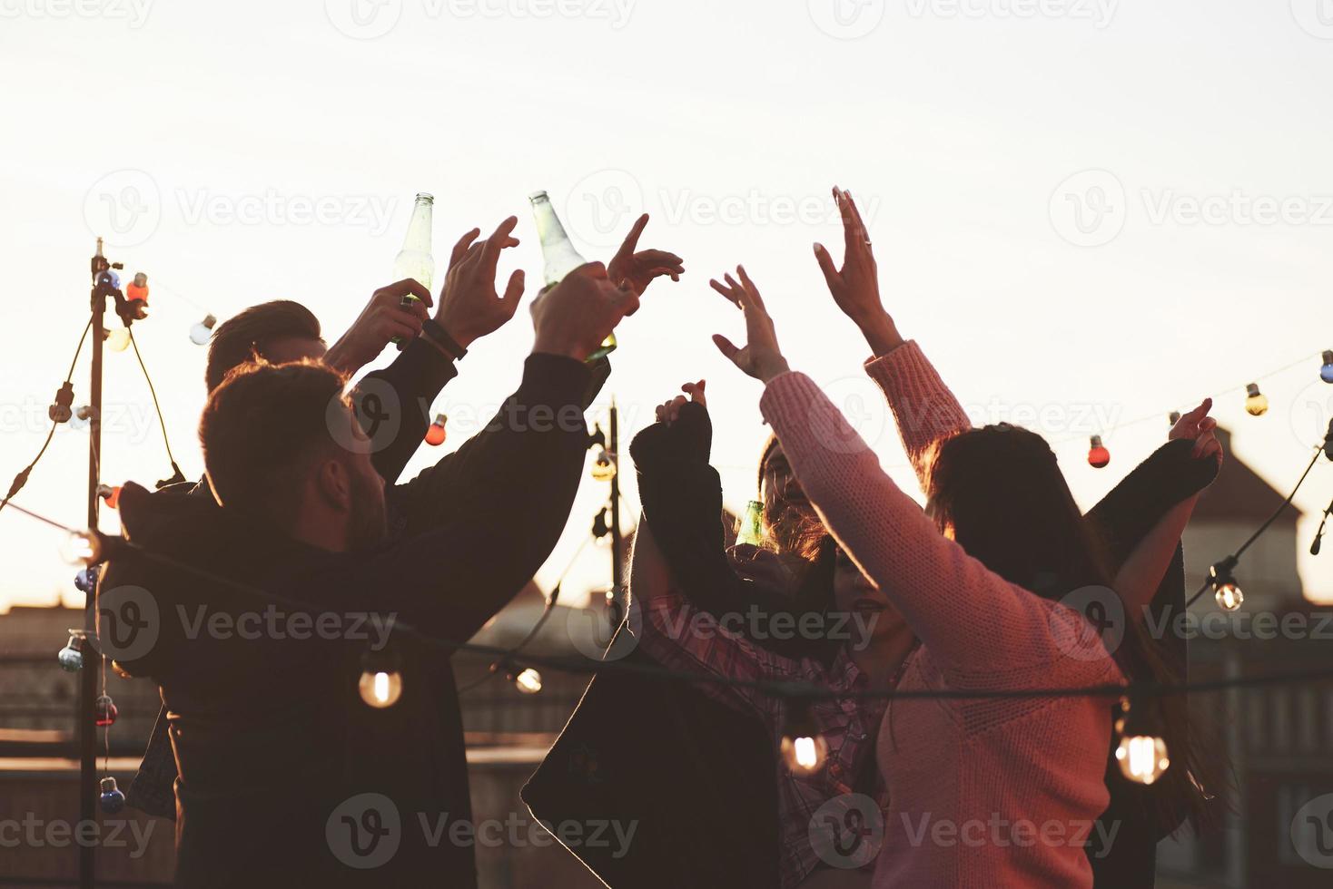 Awesome natural lighting. Holidays on the rooftop. Cheerful group of friends raised their hands up with alcohol photo