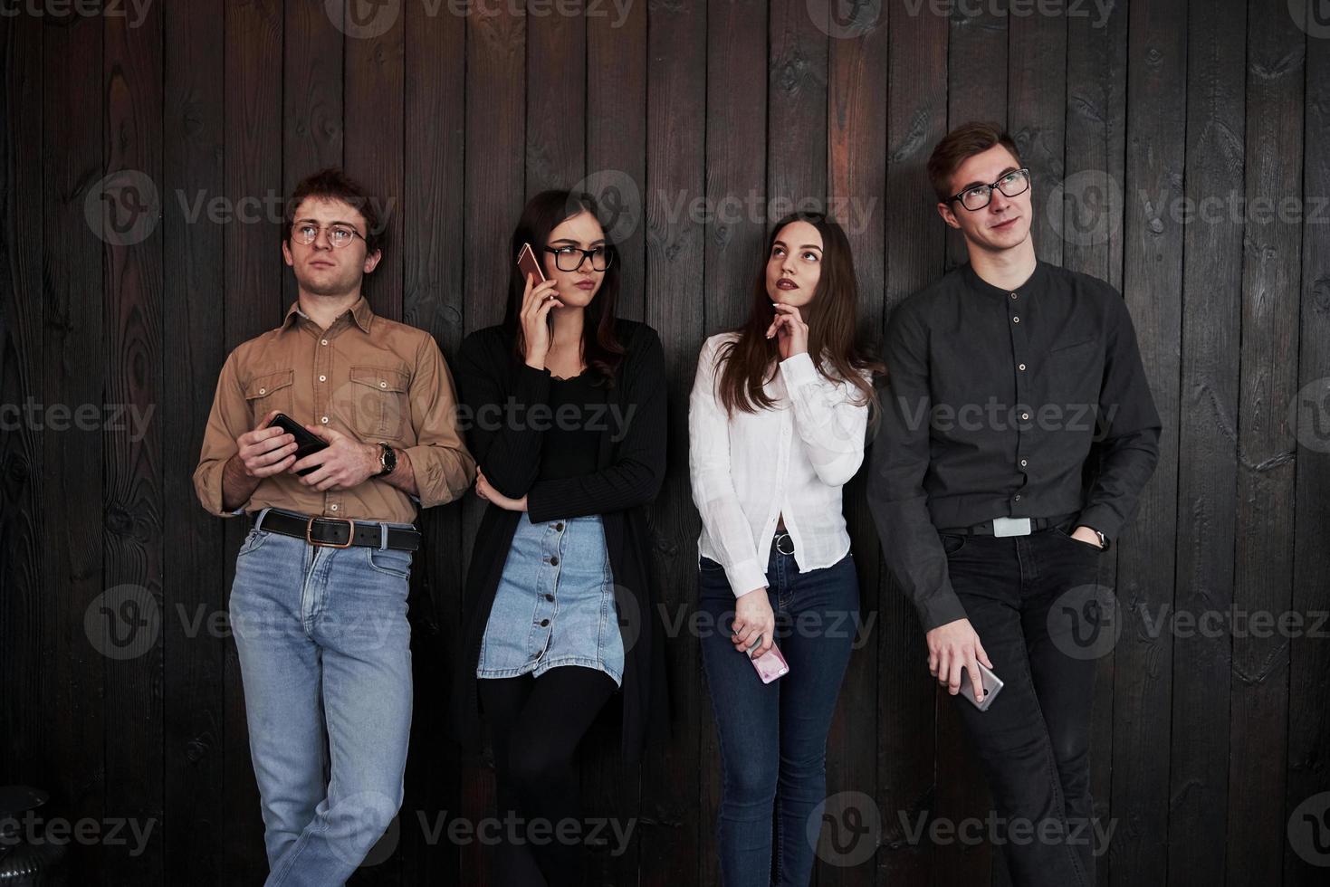 todos miran en diferentes direcciones. el joven se para contra la pared de madera negra. grupo de amigos pasando tiempo juntos foto