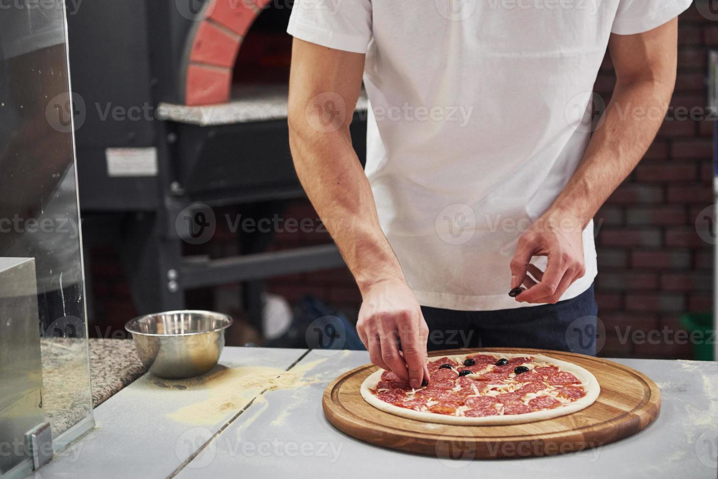 haciendo su trabajo panadero con camisa blanca poniendo aceitunas para hacer una deliciosa pizza para un pedido en el restaurante foto